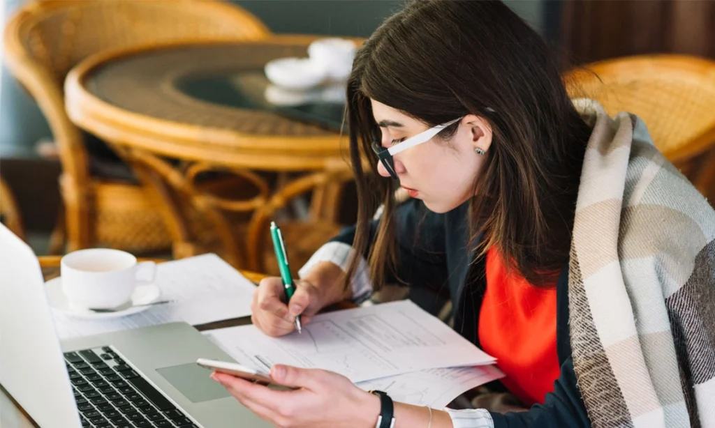Student writing a supplemental essay while reviewing college application documents on a laptop and phone